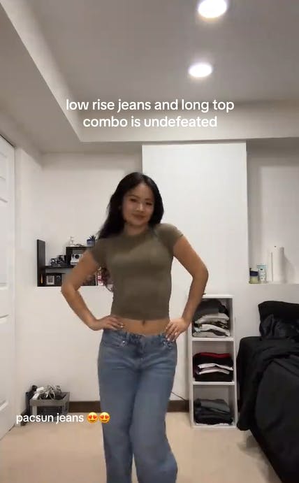 Young woman showing off her cropped t-shirt and low rise jeans. Text overlay reads, 'low rise jeans and long top combo is undefeated. pacsun jeans (heart eyes emojis)'